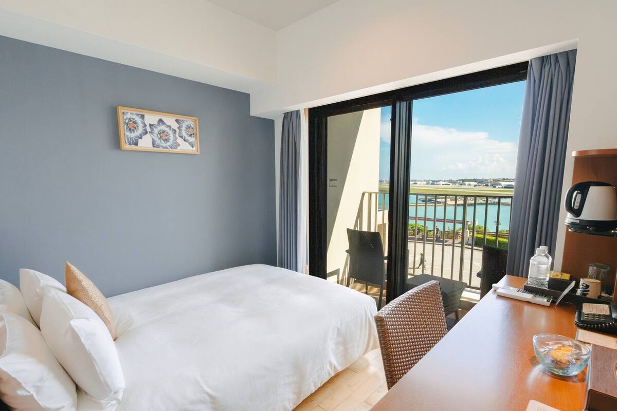 A hotel room with a bed, a desk with a laptop, a coffee maker, and a view of a body of water and a distant landscape.