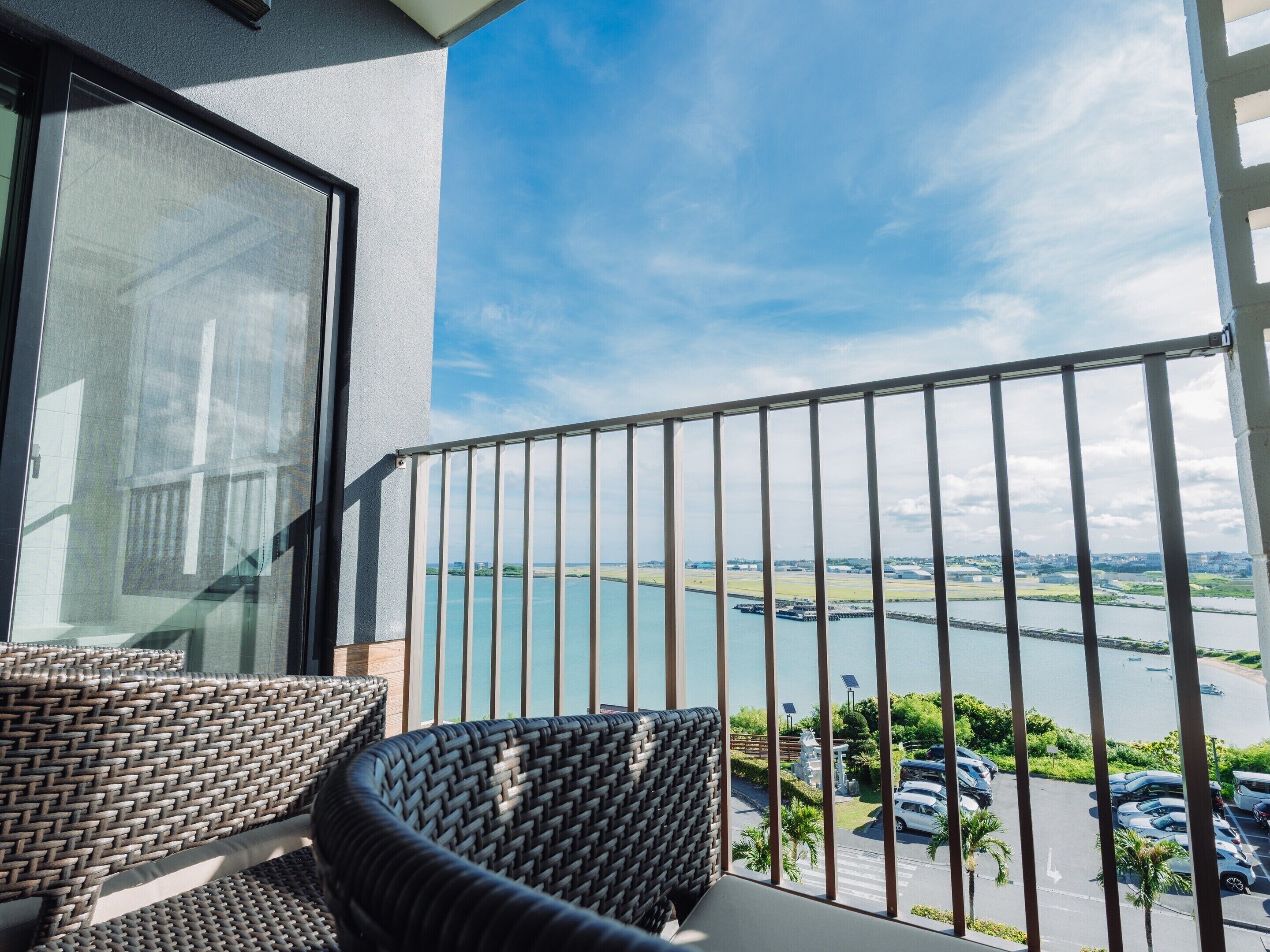 A balcony with wicker furniture overlooking a body of water and a parking area.