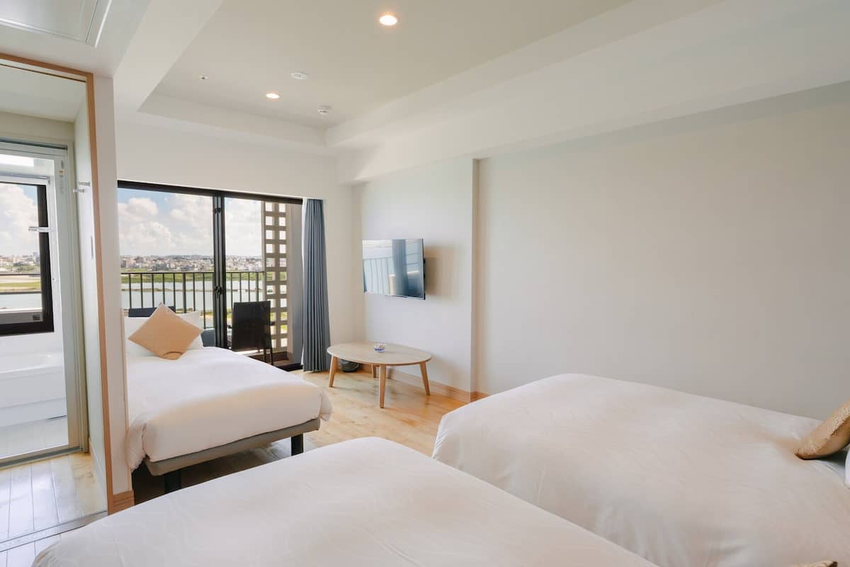 A hotel room with two beds, a small round table, a TV mounted on the wall, and a balcony with a view of the city.
