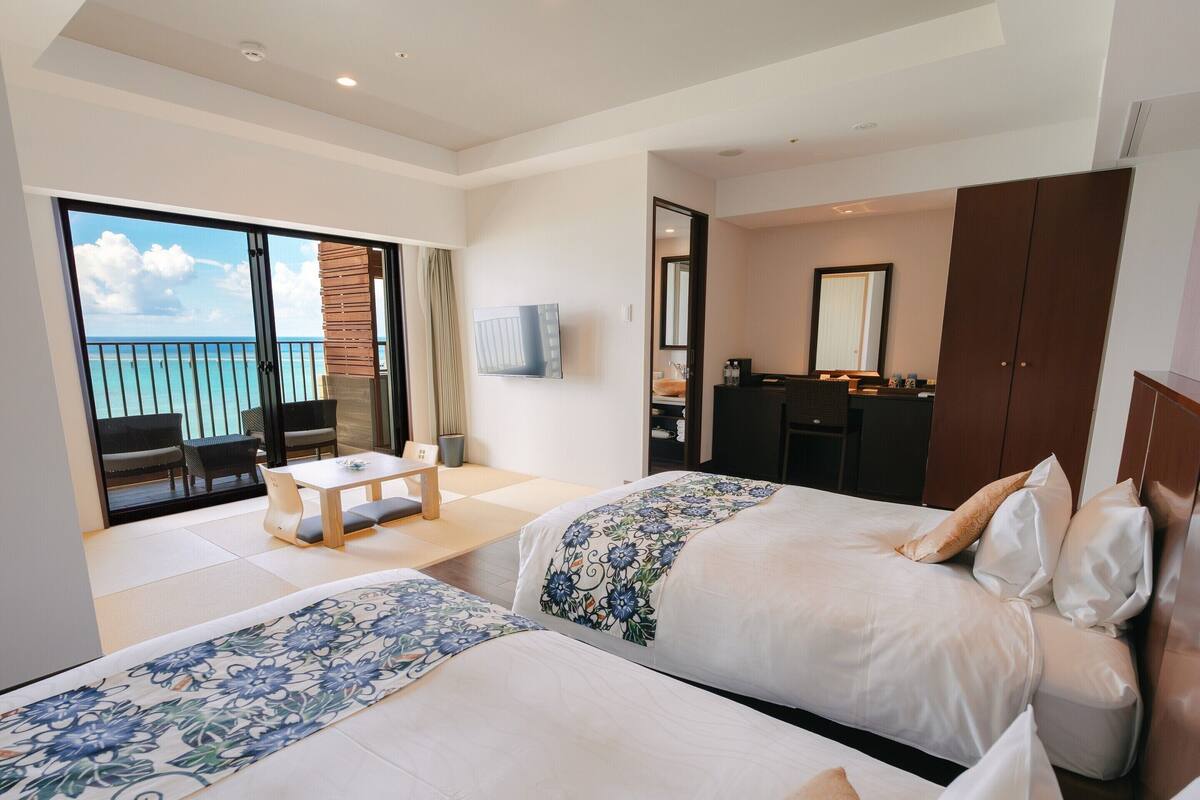 A hotel room with two beds, a desk, and a balcony with a view of the ocean.