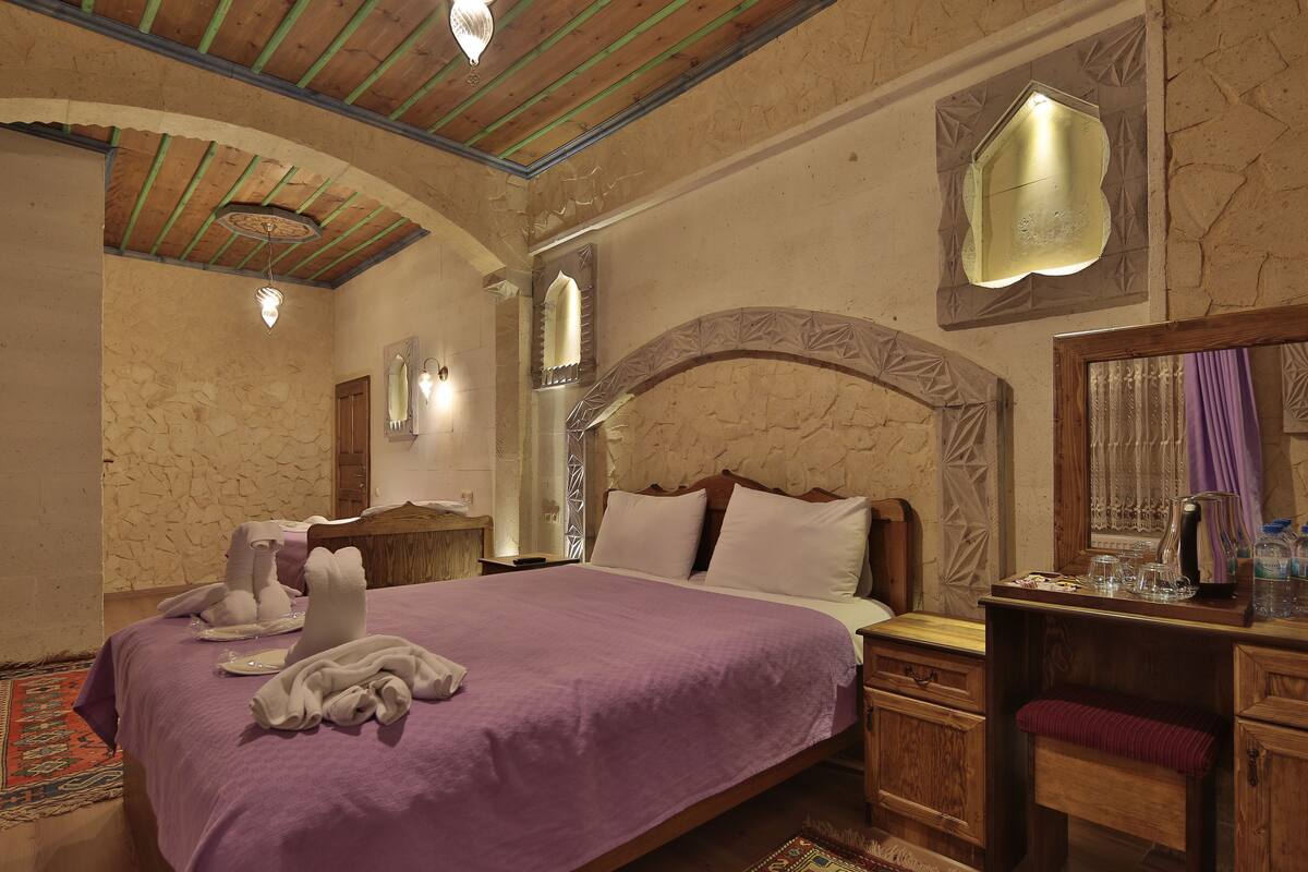 A bedroom with a stone wall, a wooden headboard, a purple bedspread, and a nightstand with a lamp and a tray of glasses.