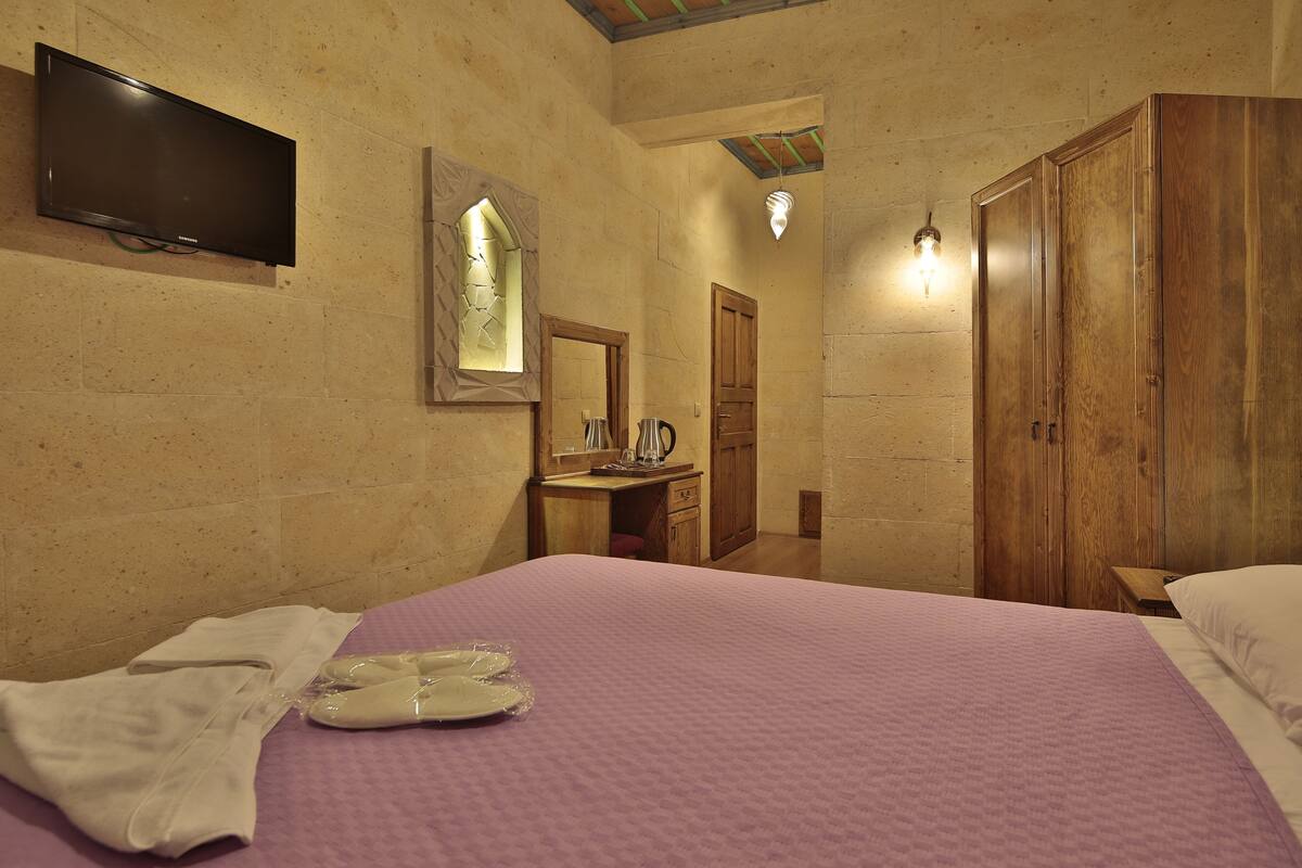 A bedroom with a bed, a television mounted on the wall, a wooden wardrobe, and a small table with a kettle.