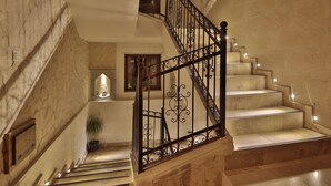 Una amplia escalera con barandillas de hierro ornamentadas y escalones de mármol, que conduce a una zona de descanso con una planta en maceta y un cuadro enmarcado.