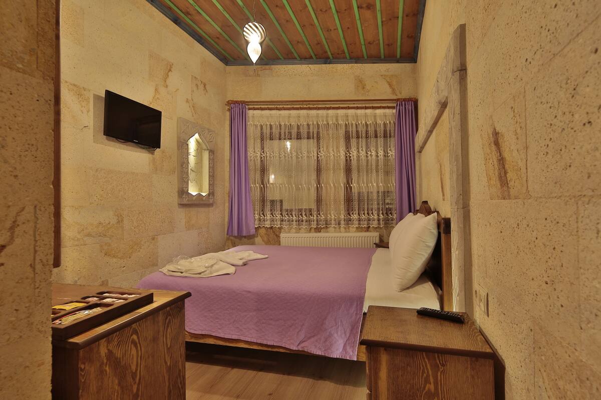 A bedroom with a bed, a TV mounted on the wall, a window with curtains, and a wooden headboard.