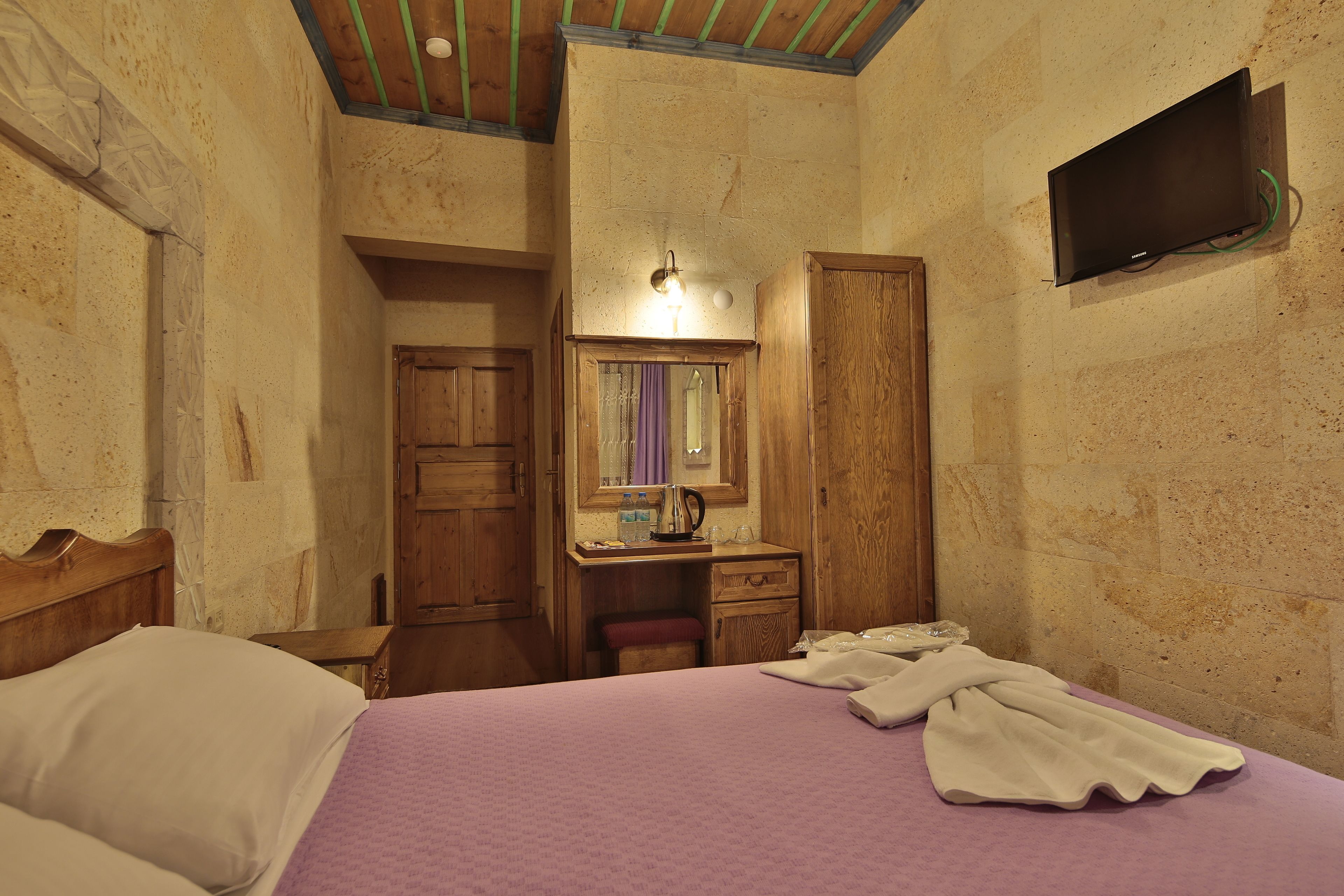 A bedroom with a bed, a wooden desk with a mirror, a television mounted on the wall, and a wooden wardrobe.