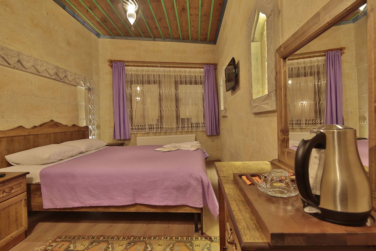 A bedroom with a wooden bed, purple curtains, a wooden nightstand, a bedside table with a kettle and a glass, and a patterned rug.