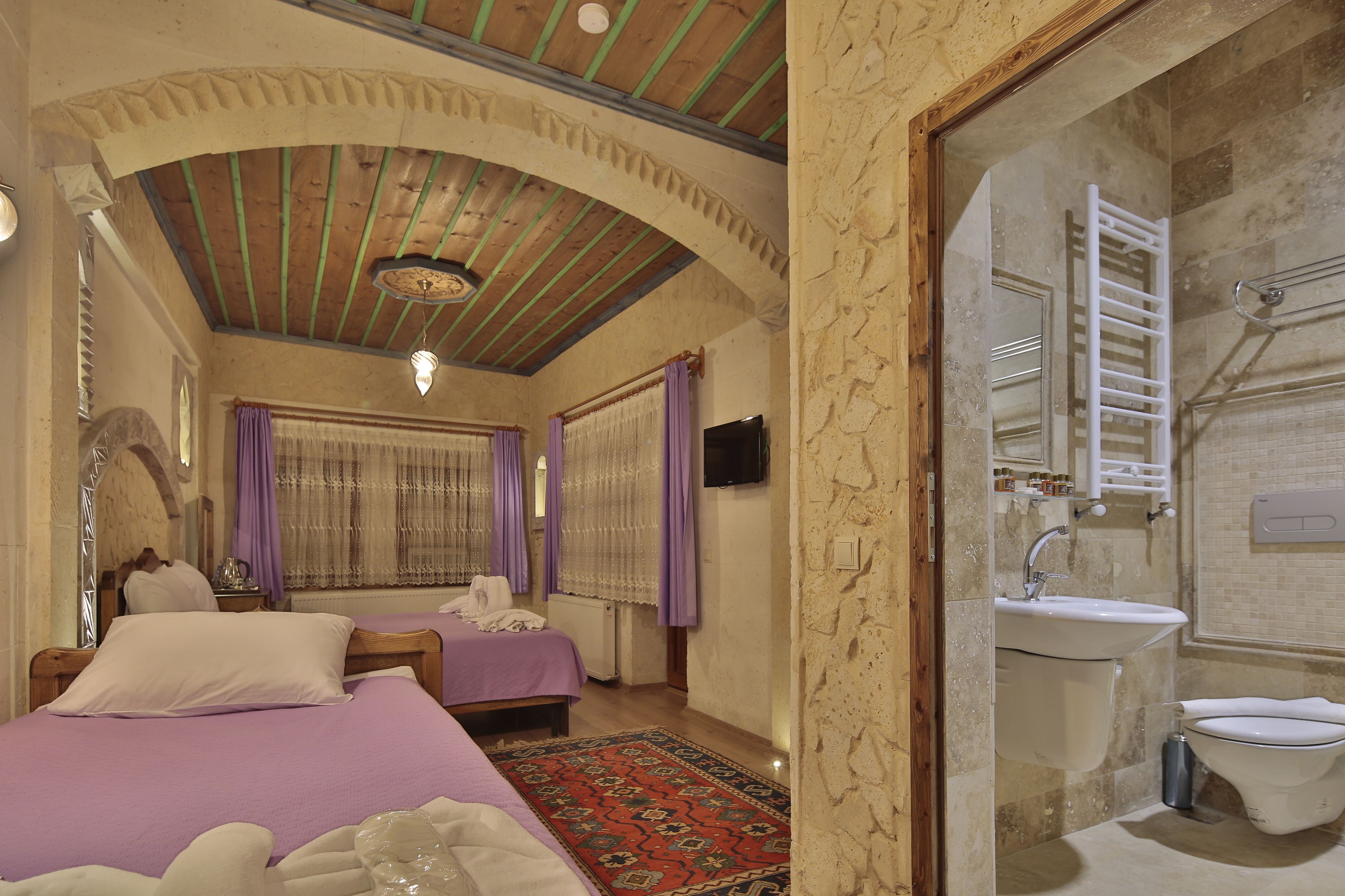 A room with two beds, a red patterned rug, a wooden ceiling, and a bathroom with a sink and toilet.