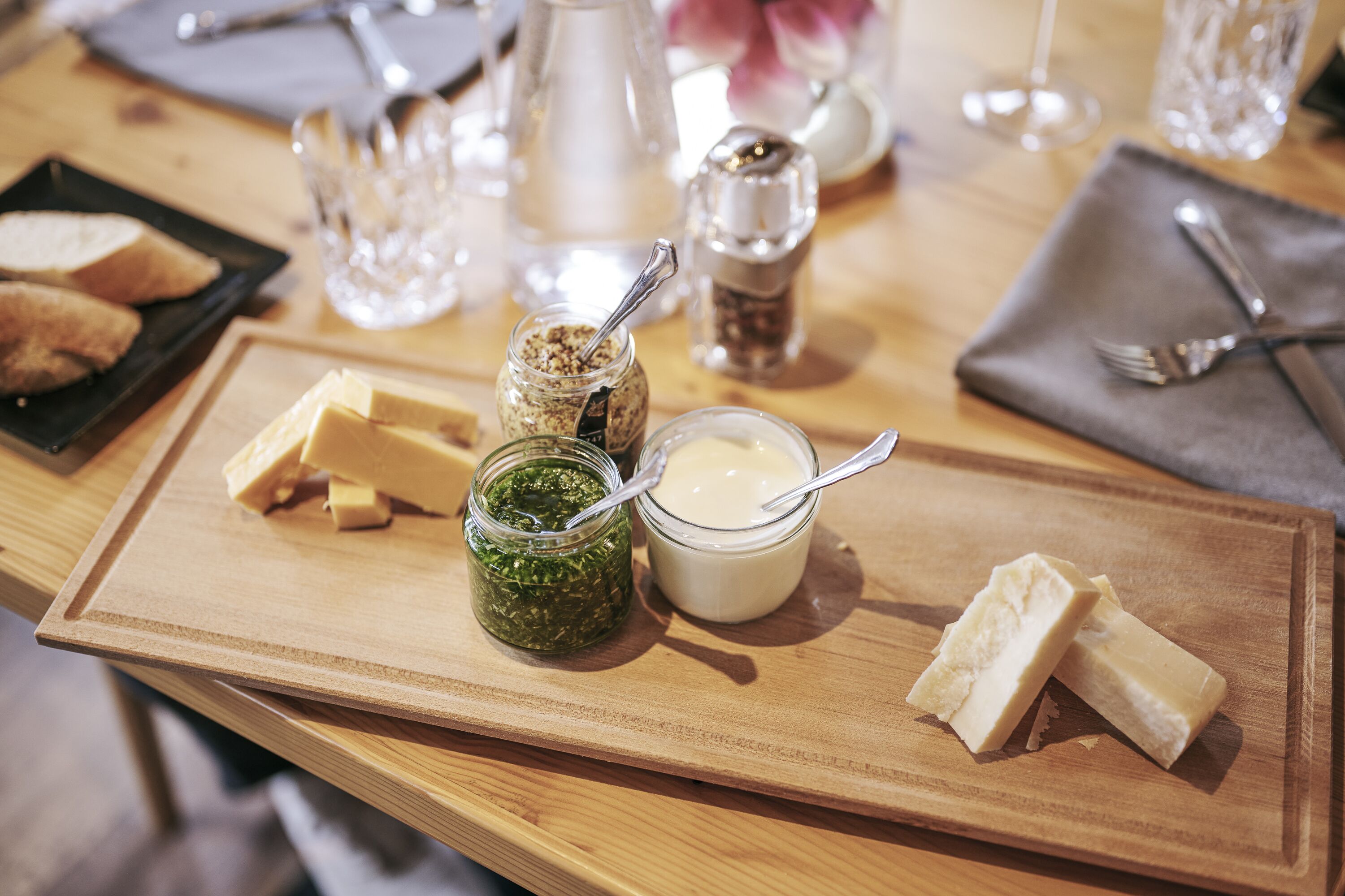 A wooden serving board with cheese, a jar of green spread, and a bowl of cream on a table.