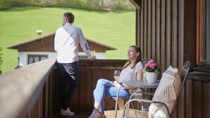 A balcony with a wooden railing, a table with a potted plant, and a view of a green landscape. - Der Klostertalerhof (Klösterle am Arlberg)