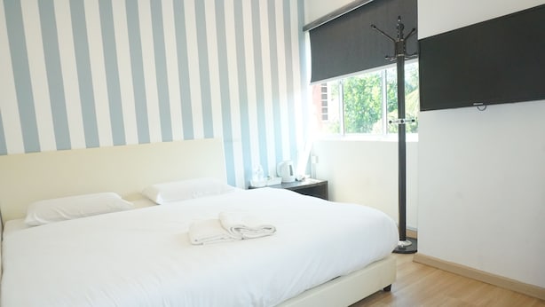 Una cama bien hecha con ropa blanca y una toalla doblada, una mesita de noche con lámpara, y una ventana con vista a follaje exterior.