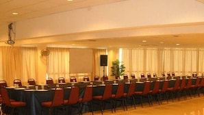 Un salón de conferencias con mesas largas y sillas, un proyector y cortinas.