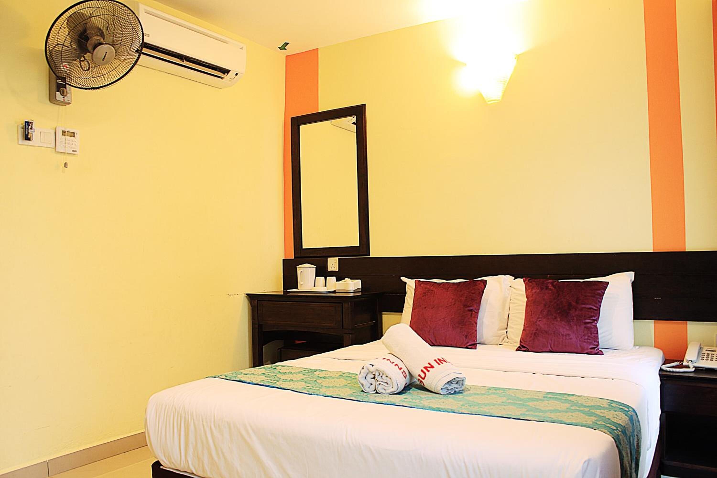 A hotel room with a bed, a bedside table, a nightstand, a wall-mounted air conditioner, a wall-mounted mirror, and a wall-mounted light fixture.