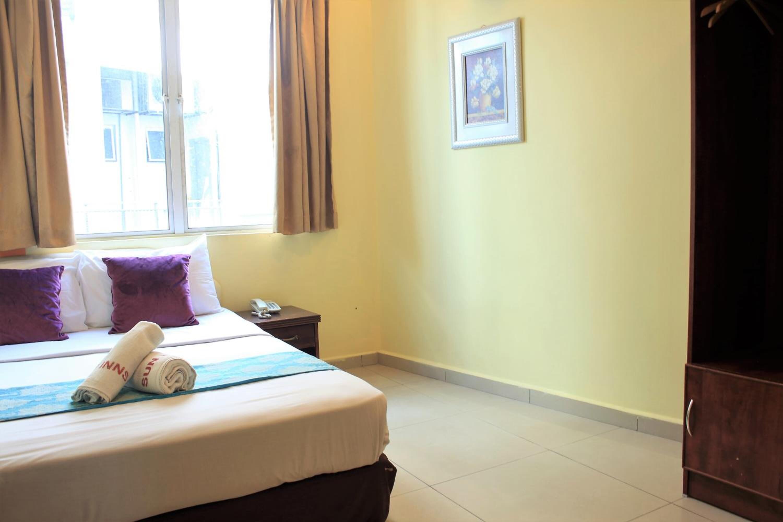 Quarto de hotel com cama, mesa de cabeceira, guarda-roupa, janela com cortinas e um quadro na parede.