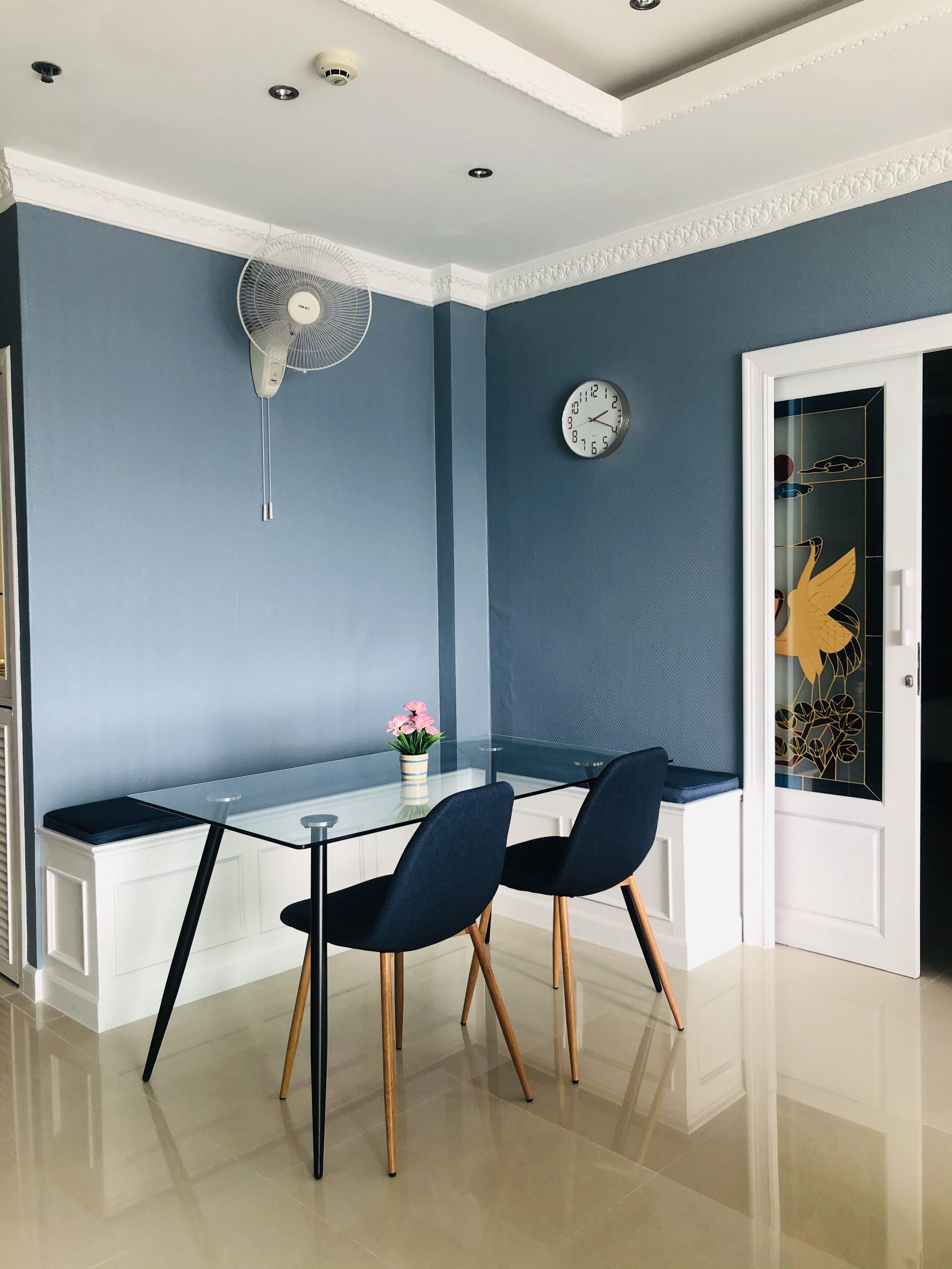A modern dining area with a glass table, two chairs, a wall-mounted fan, and a clock.