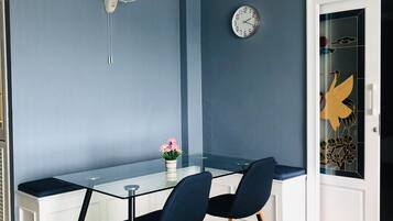 A modern dining area with a glass table, two chairs, a wall-mounted fan, and a clock.