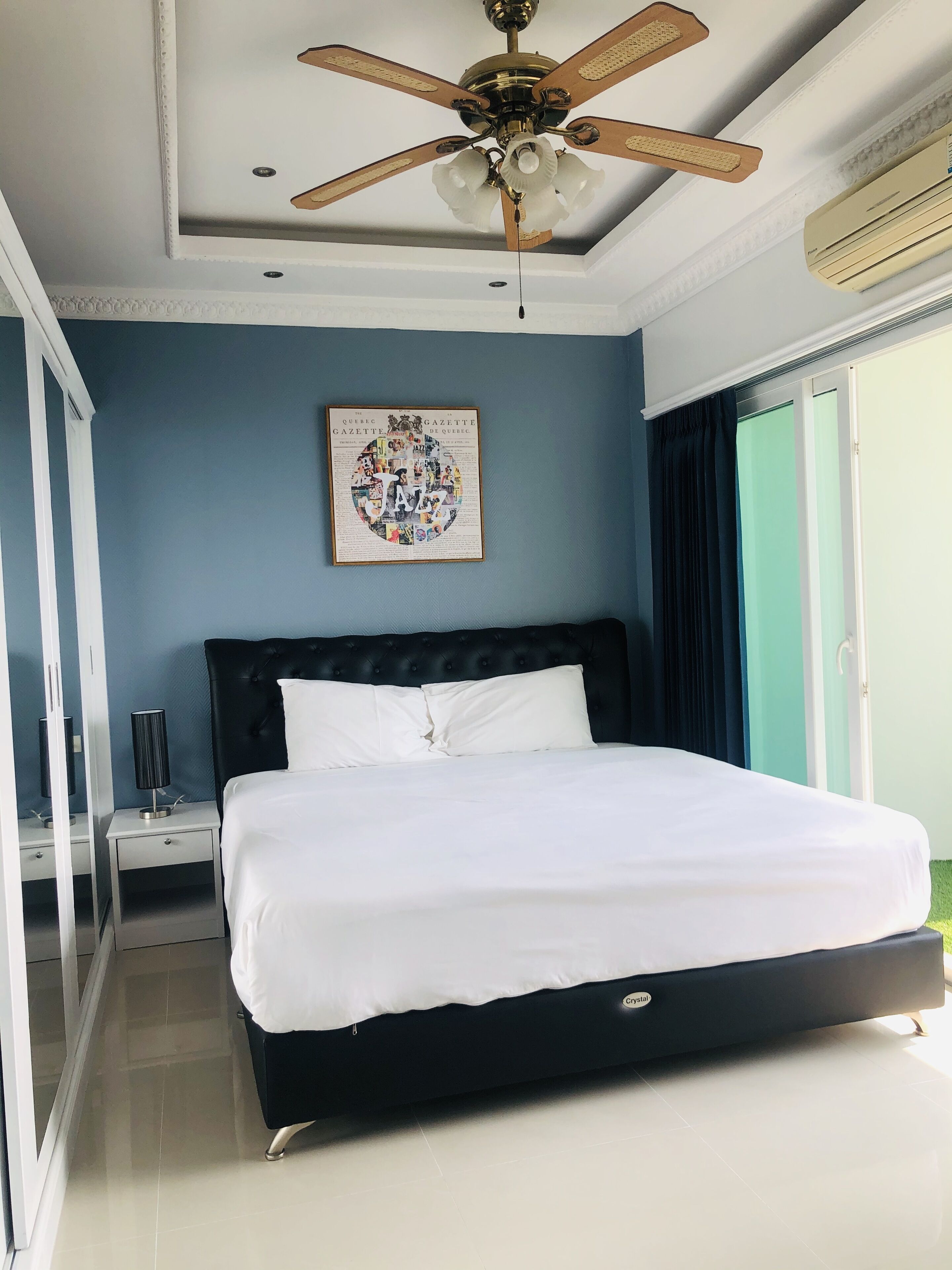 A bedroom with a large bed, a ceiling fan, a framed picture on the wall, and a window with curtains.