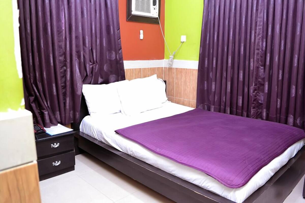 A neatly made bed with white linens and a purple blanket, a bedside table with a lamp, and a wall-mounted air conditioner.