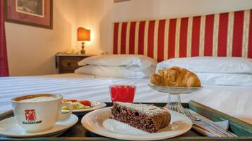 Ein Hotelzimmer mit einem Bett, einem Nachttisch mit Lampe, einem Tablett mit Frühstücksangeboten, darunter ein Stück Kuchen, ein Glas roter Saft und ein Croissant.