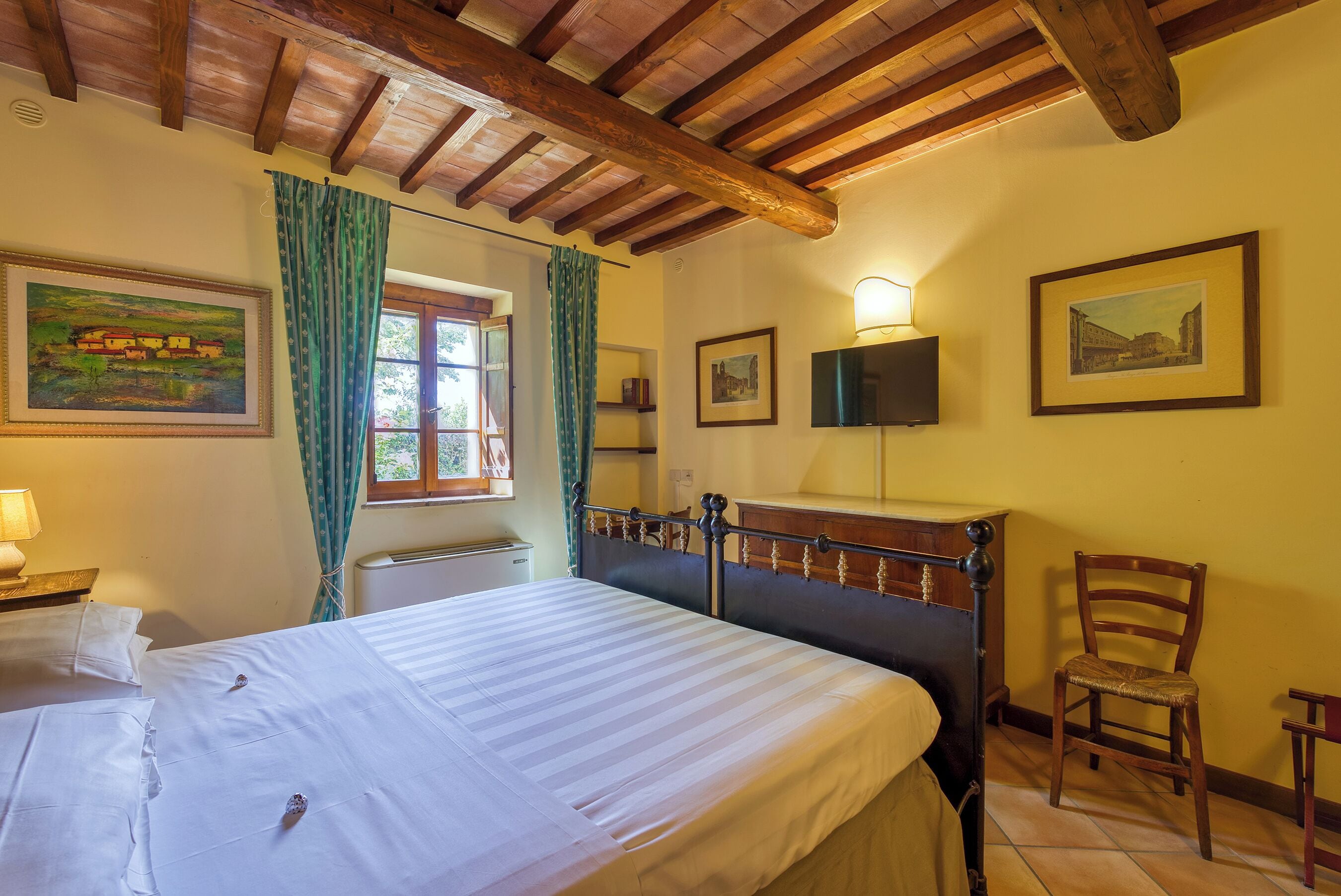 Ein Schlafzimmer mit Holzbalkendecke, einem Bett mit weißer Bettwäsche, einem Sessel und zwei gerahmten Bildern an der Wand.
