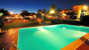 A swimming pool with a clear blue water surface, surrounded by a well-lit patio area with lounge chairs and a brick pathway. - Borgo Mandoleto (Perugia)