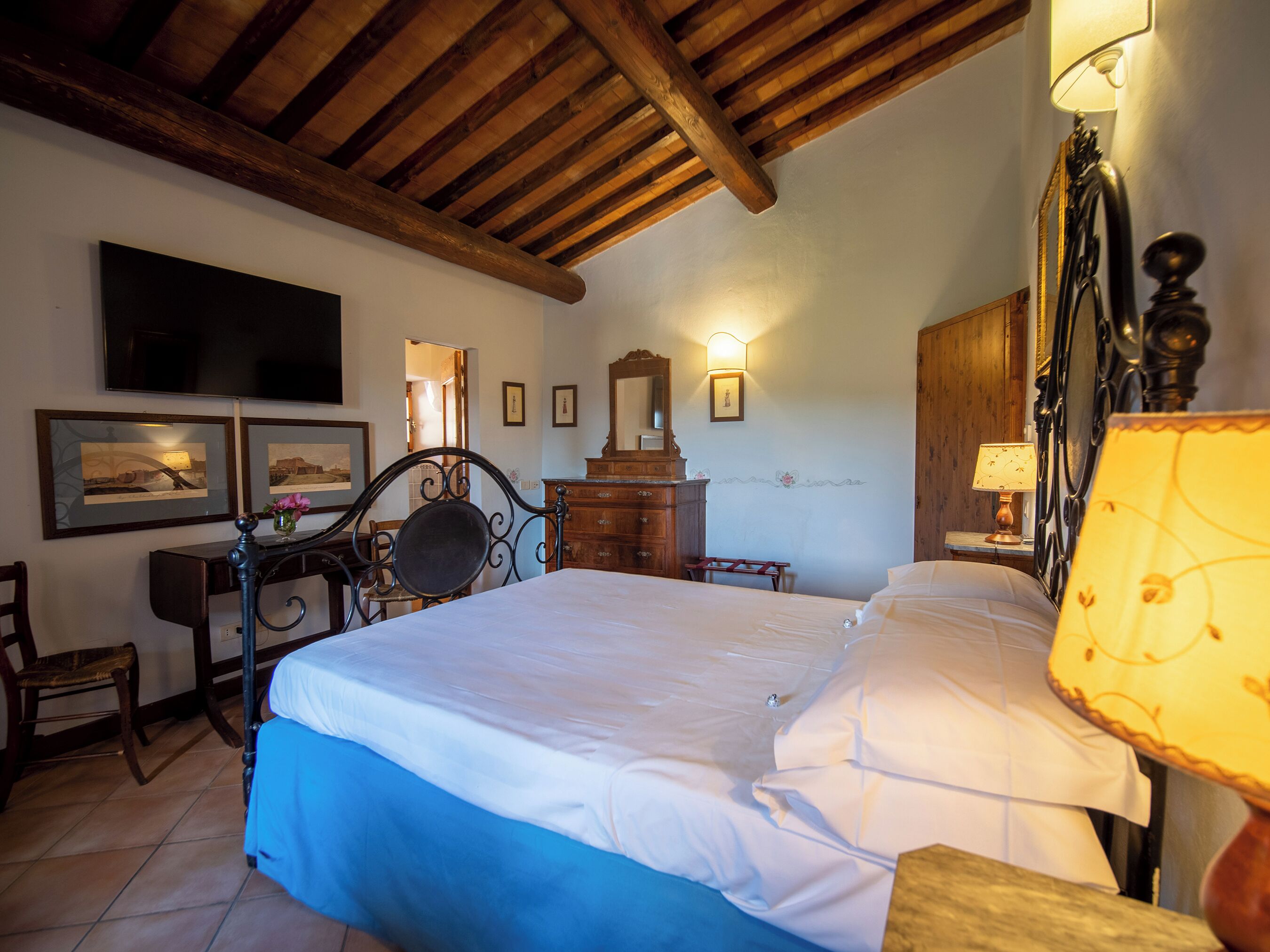 A bedroom with a wooden ceiling, a bed with white bedding, a desk with a chair, a dresser, and wall-mounted lamps.
