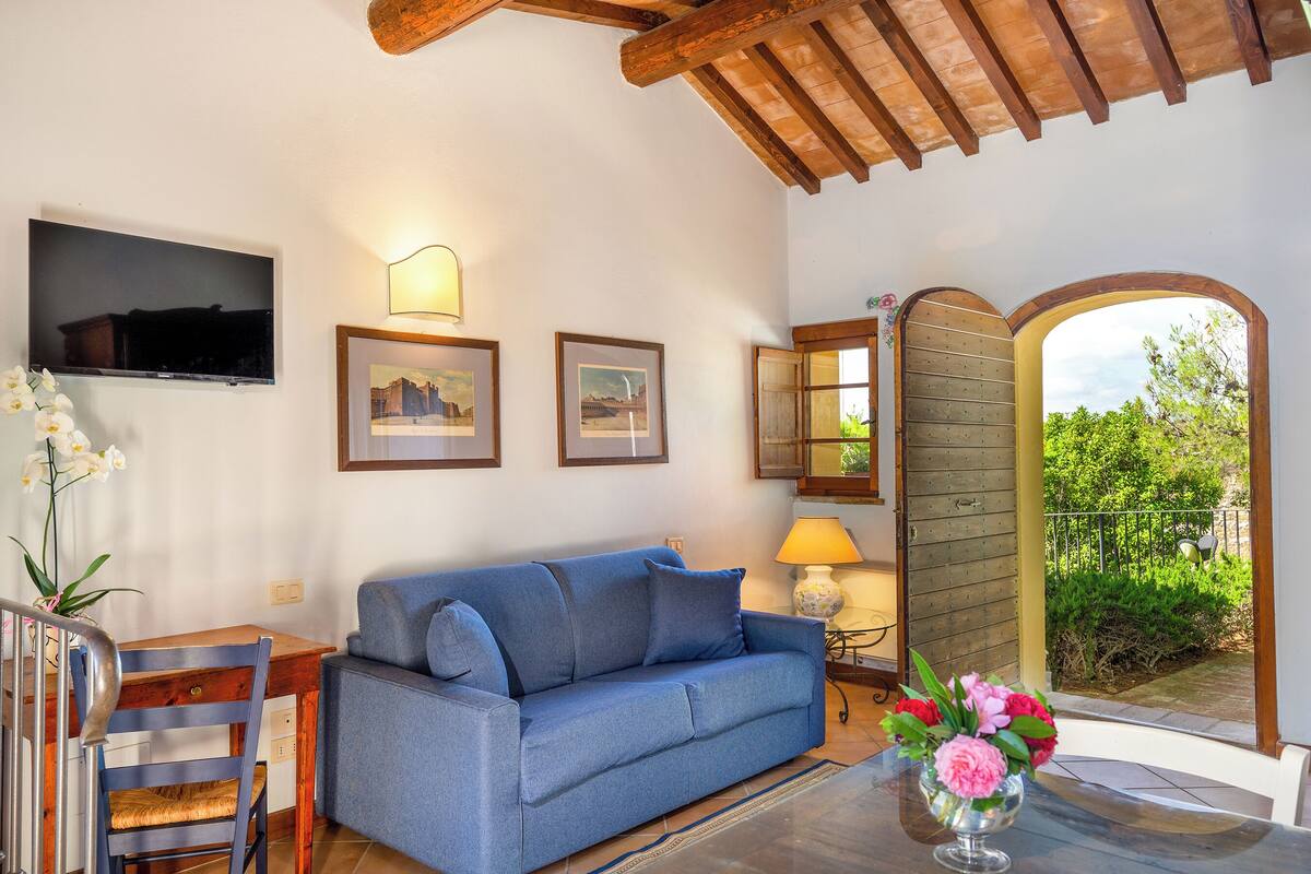 A cozy living area with a blue sofa, wooden dining table, and chairs. There are framed pictures on the wall, a TV mounted above the sofa, and a window with a view of greenery.