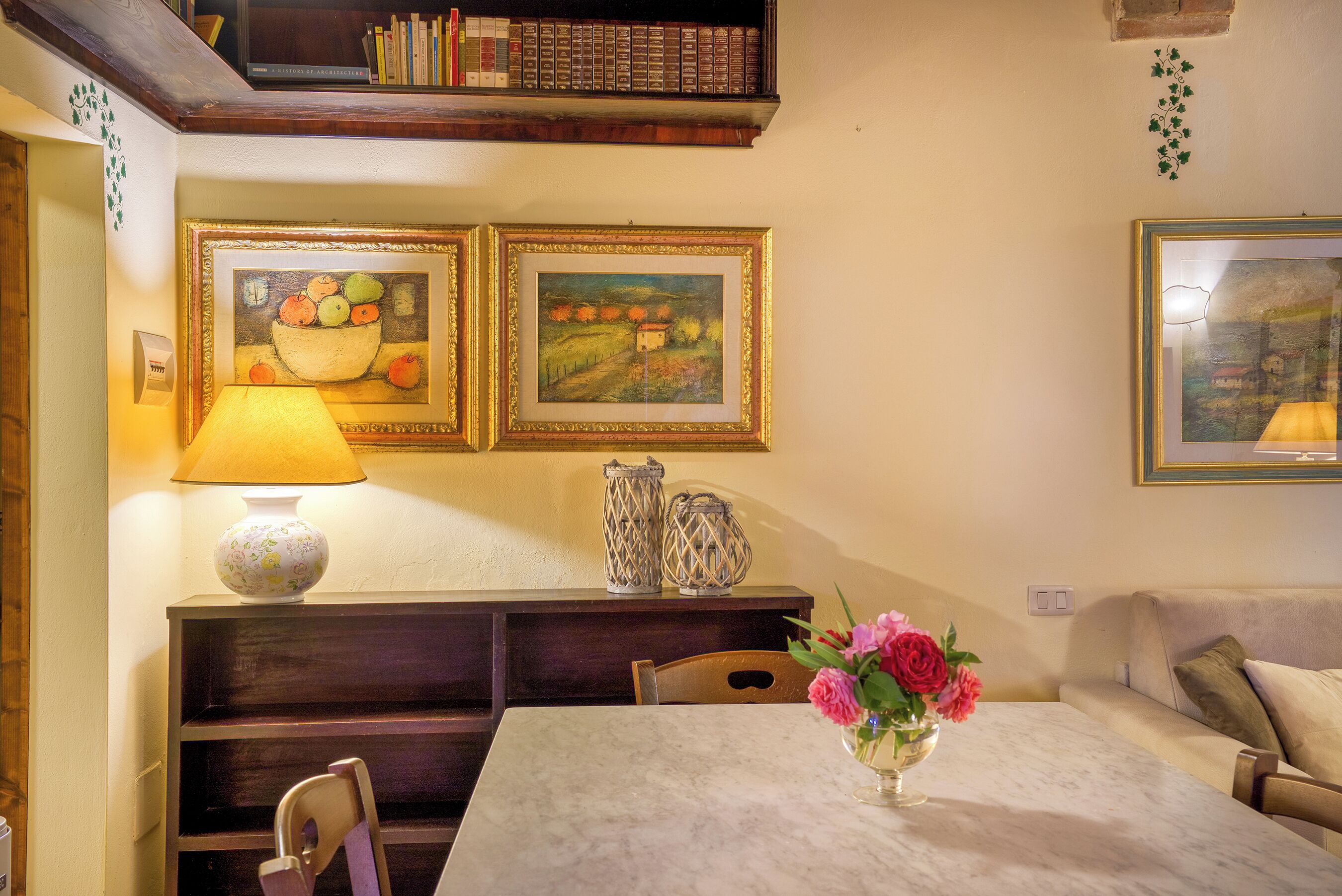 A dining area with a table, chairs, a lamp, and two framed paintings on the wall.
