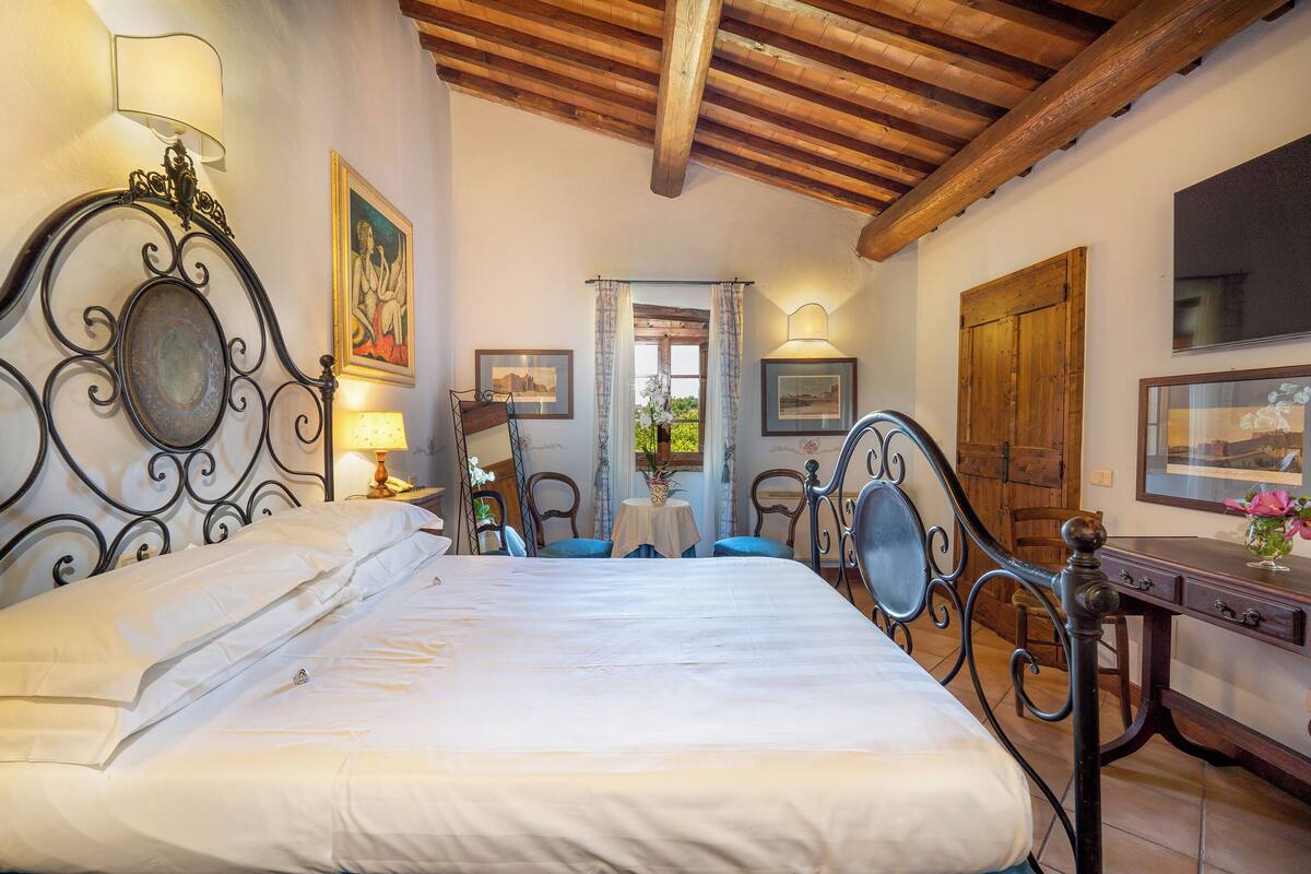 A bedroom with a large bed, a wooden ceiling, a desk with a lamp, and a painting on the wall.