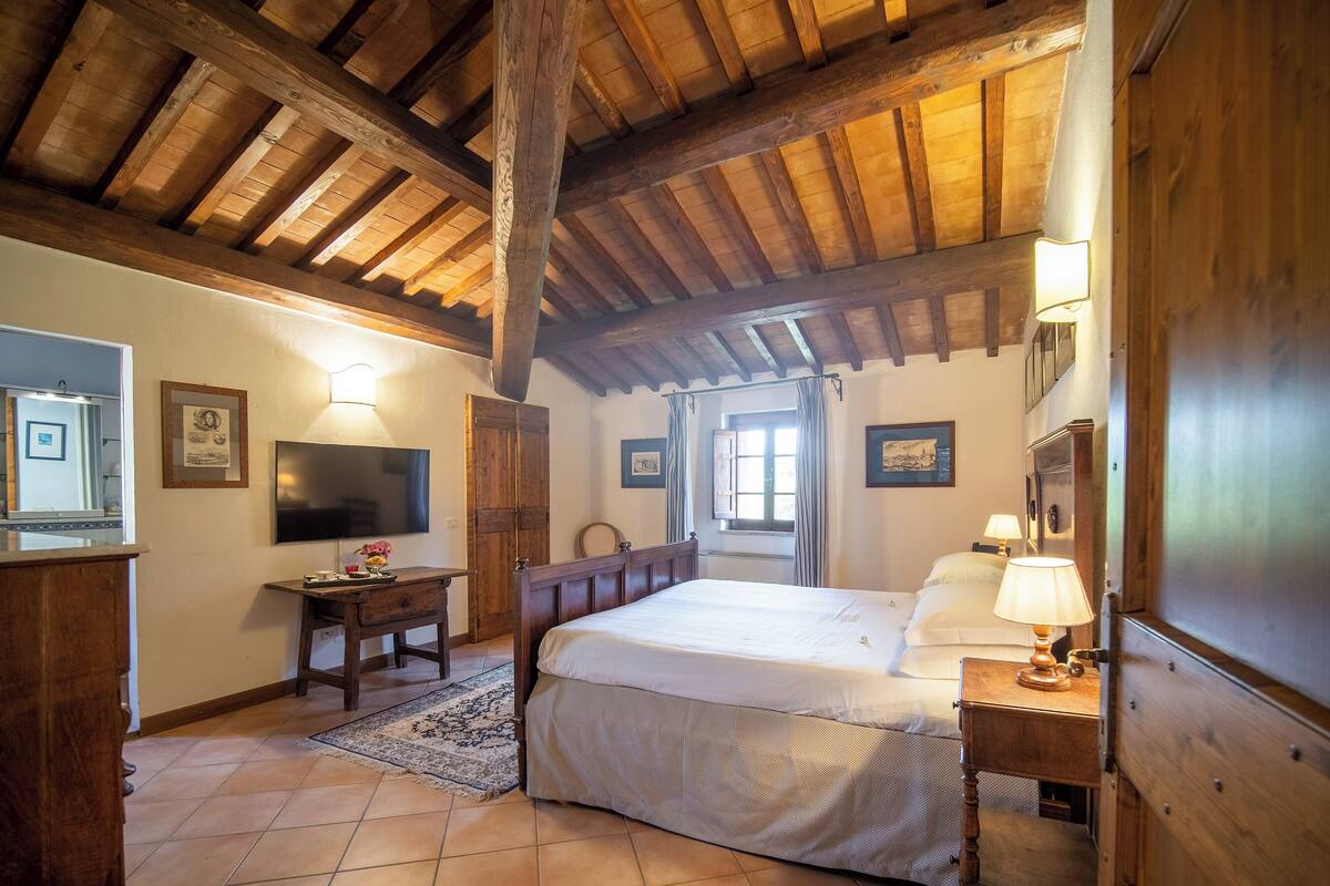 A bedroom with a wooden ceiling, a bed with white linens, a desk with a lamp, and a TV on the wall.