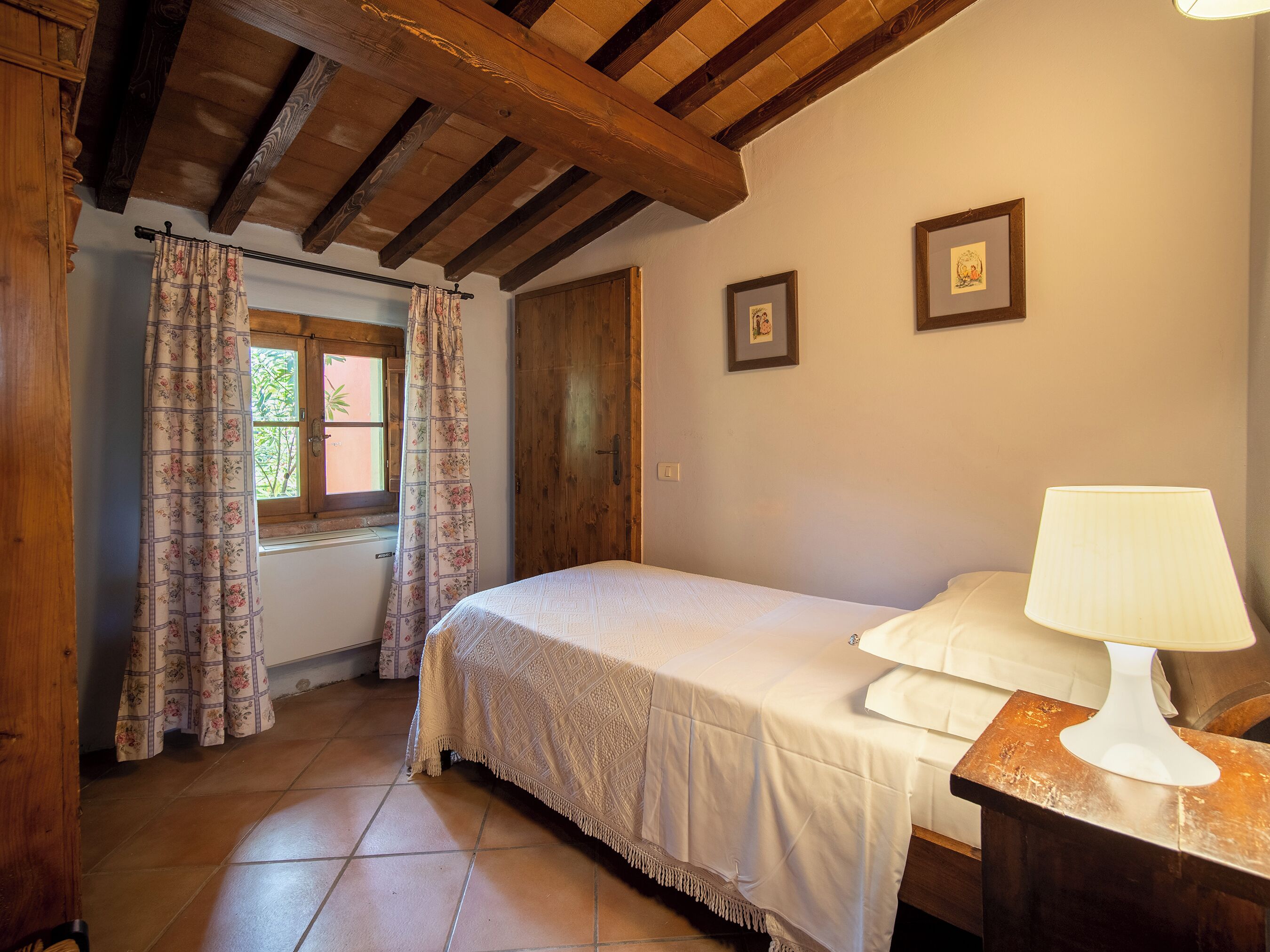 Quarto com cama, mesa de cabeceira, luminária, vigas de madeira, janela com cortinas e porta.