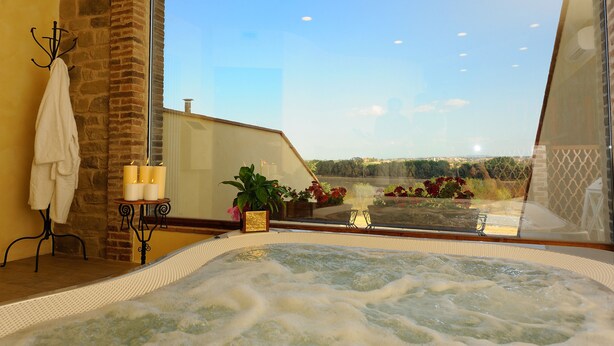 A spa room with a hot tub, a stone wall, a window with a view of the countryside, and a small table with candles. - Borgo Mandoleto (Perugia)