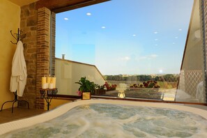 A spa room with a hot tub, a stone wall, a window with a view of the countryside, and a small table with candles. - Borgo Mandoleto (Perugia)
