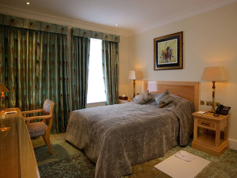 A hotel room with a large bed, a chair, a desk, and a nightstand with a lamp.