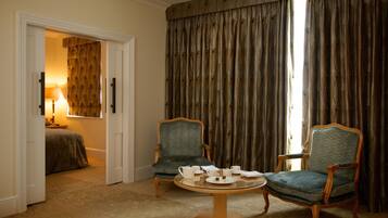 Une chambre d’hôtel comprenant un lit, deux fauteuils, une petite table ronde avec un service à thé et une grande fenêtre avec des rideaux.