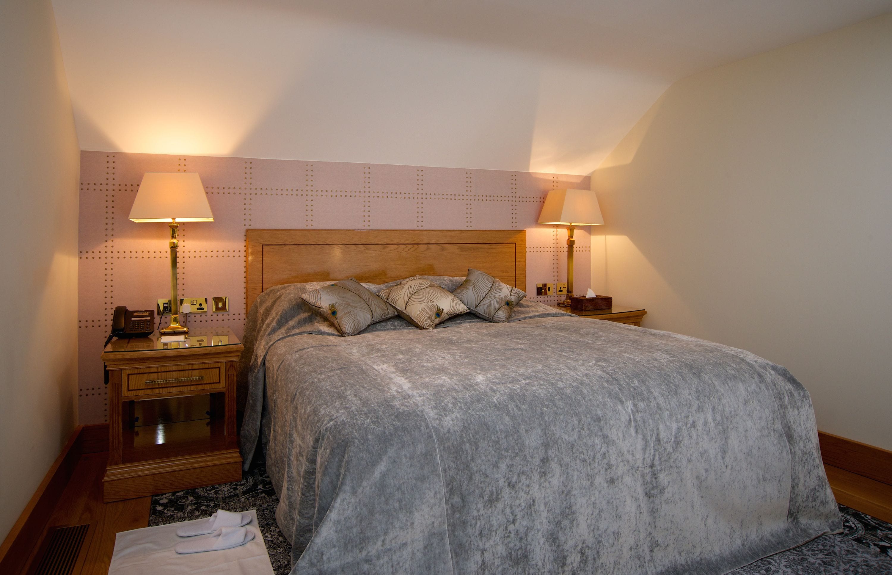 A neatly made bed with a grey blanket and decorative pillows, two bedside lamps, a wooden nightstand with a telephone and a small table, and a patterned rug on the floor.