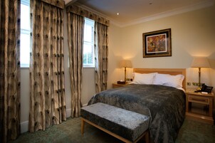 A neatly arranged bedroom with a large bed, a bedside table with a lamp, a bench, and a window with curtains.