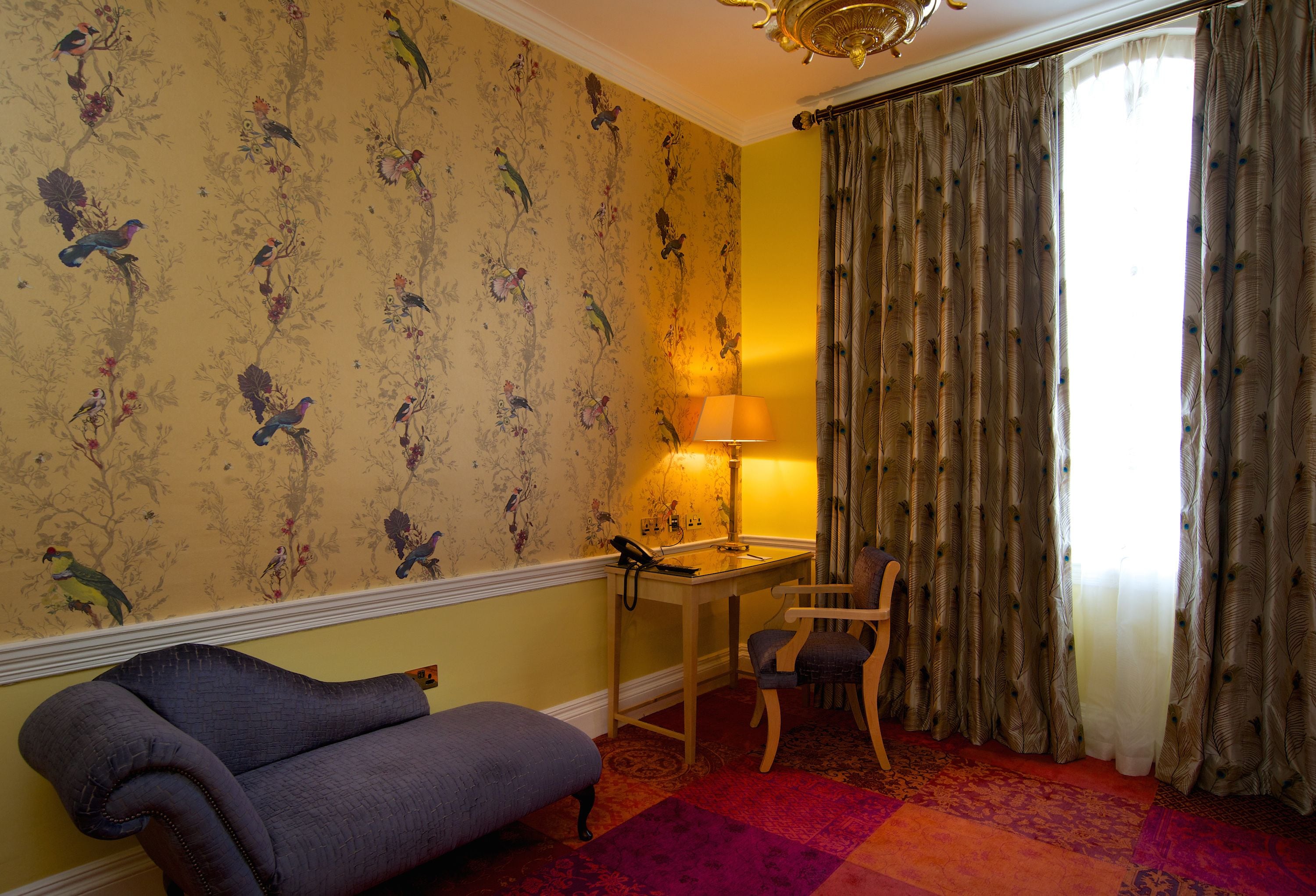 A room with a floral wallpaper, a desk with a lamp, a chair, and a sofa.
