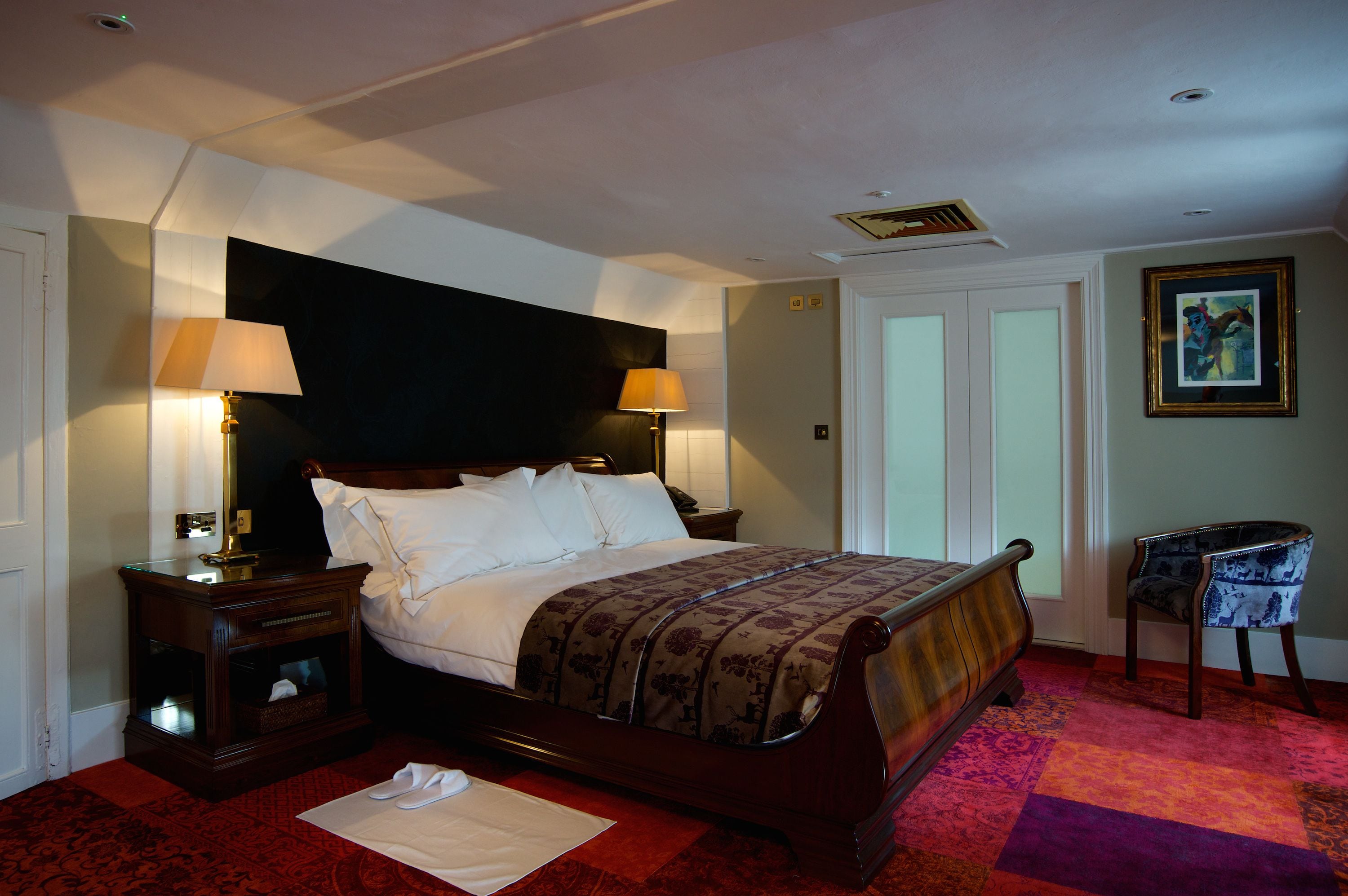 A neatly made bed with a patterned bedspread, two bedside tables with lamps, a chair, and a framed picture on the wall.