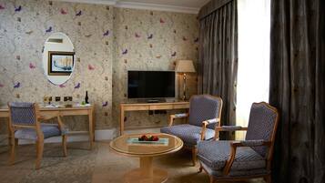 A hotel room with a flat-screen TV, two patterned armchairs, a wooden coffee table, a desk with a lamp, and a large window with curtains.