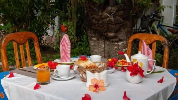 Une table dressée pour deux personnes, avec une nappe blanche, des serviettes rayées de rouge et de blanc, une théière blanche et un verre de jus d’orange.