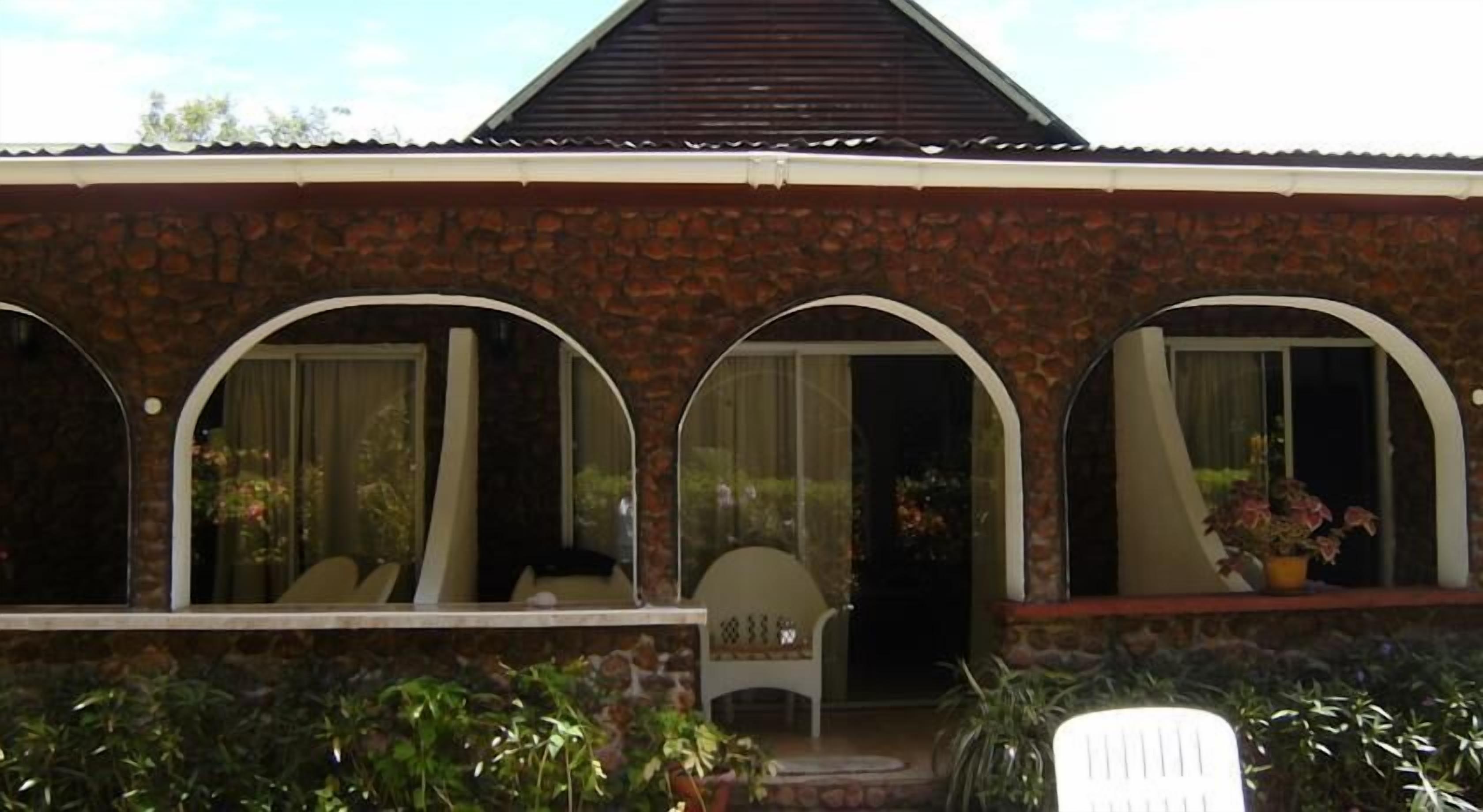 Una casa con fachada de piedra, tres ventanas en arco y un pequeño porche con una silla y plantas en macetas.