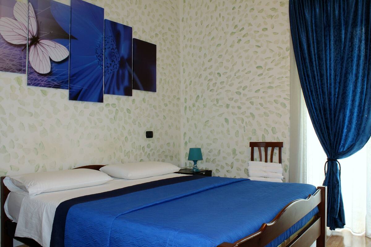 A neatly made bed with a blue bedspread, white pillows, and a wooden headboard. A blue lamp is on a bedside table, and a blue curtain is drawn to the side.