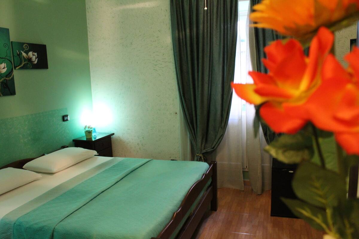A hotel room with a bed, a nightstand, a vase with flowers, and a painting on the wall.
