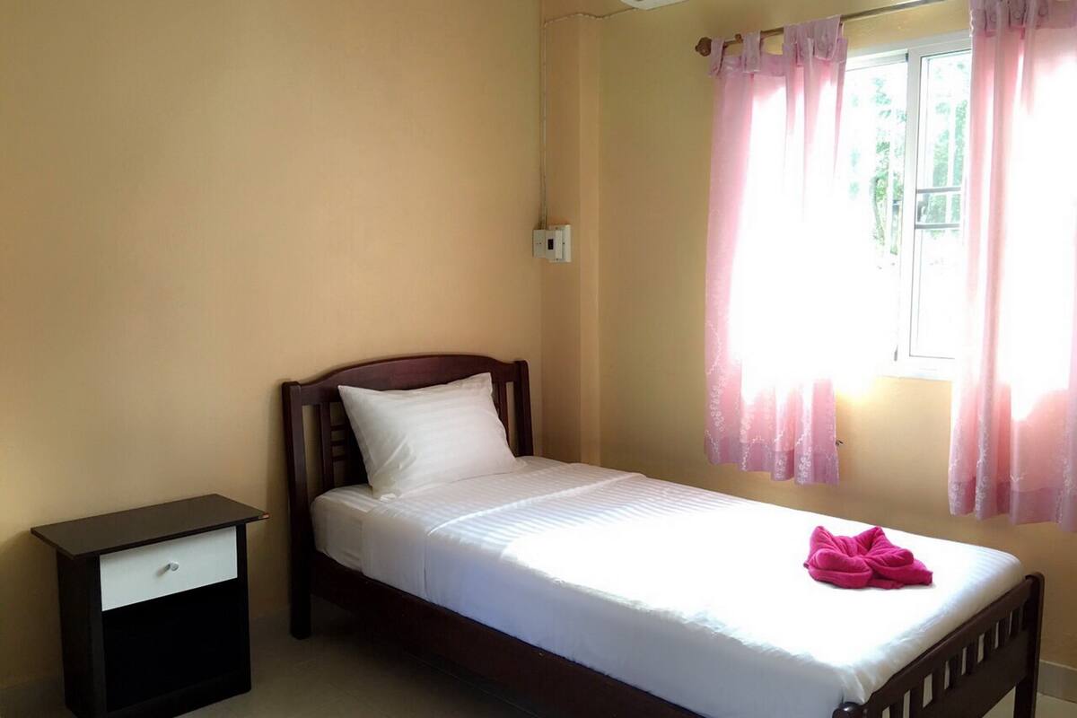 A single bed with a wooden frame and white bedding, a small bedside table with a drawer, and a window with pink curtains.