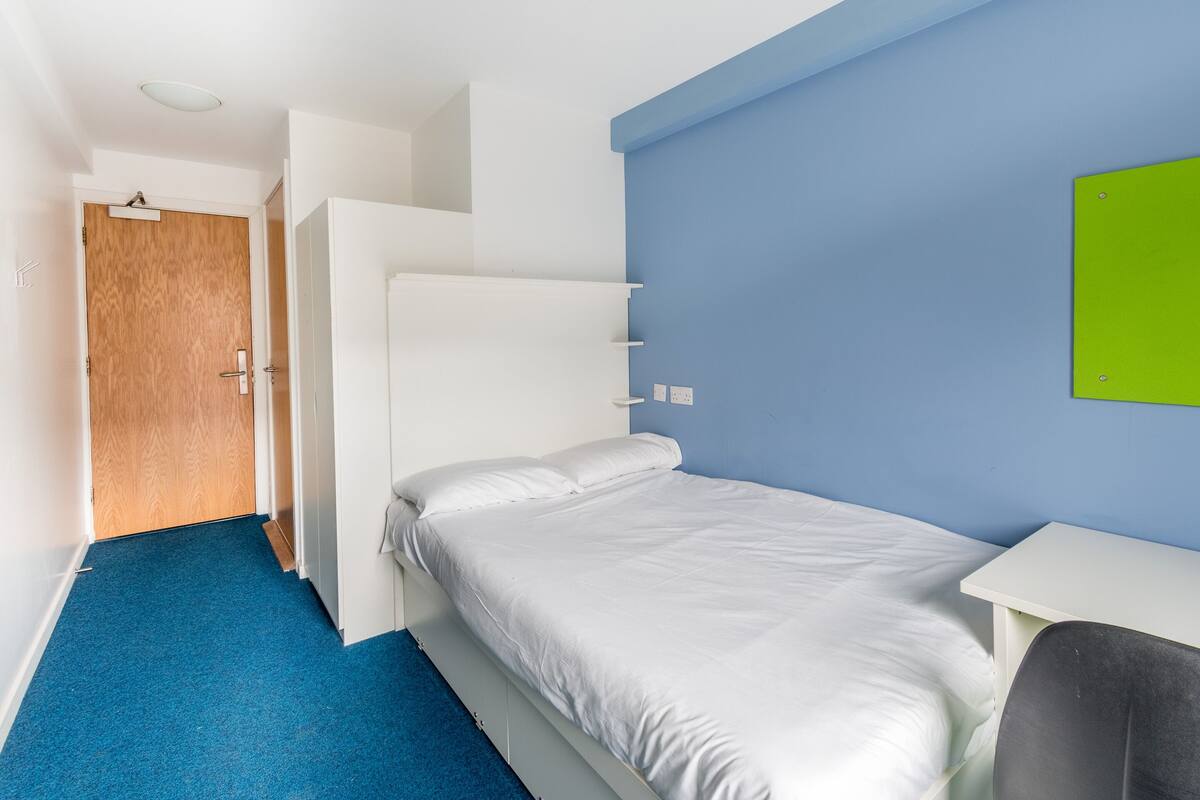 Quarto de hotel com cama, escrivaninha, cadeira, guarda-roupa e porta.