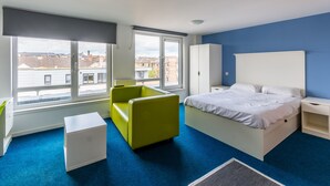 Un dormitorio moderno con una cama grande, una silla verde, una mesita de noche blanca y vistas a edificios a través de amplias ventanas.