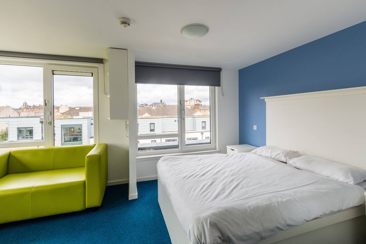 Um quarto com uma cama, um sofá verde e uma janela com vista para edifícios.