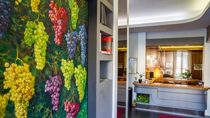 A reception area with a modern desk, a large wall painting of grapes, and a red carpet. - Hotel Italia (Stradella)