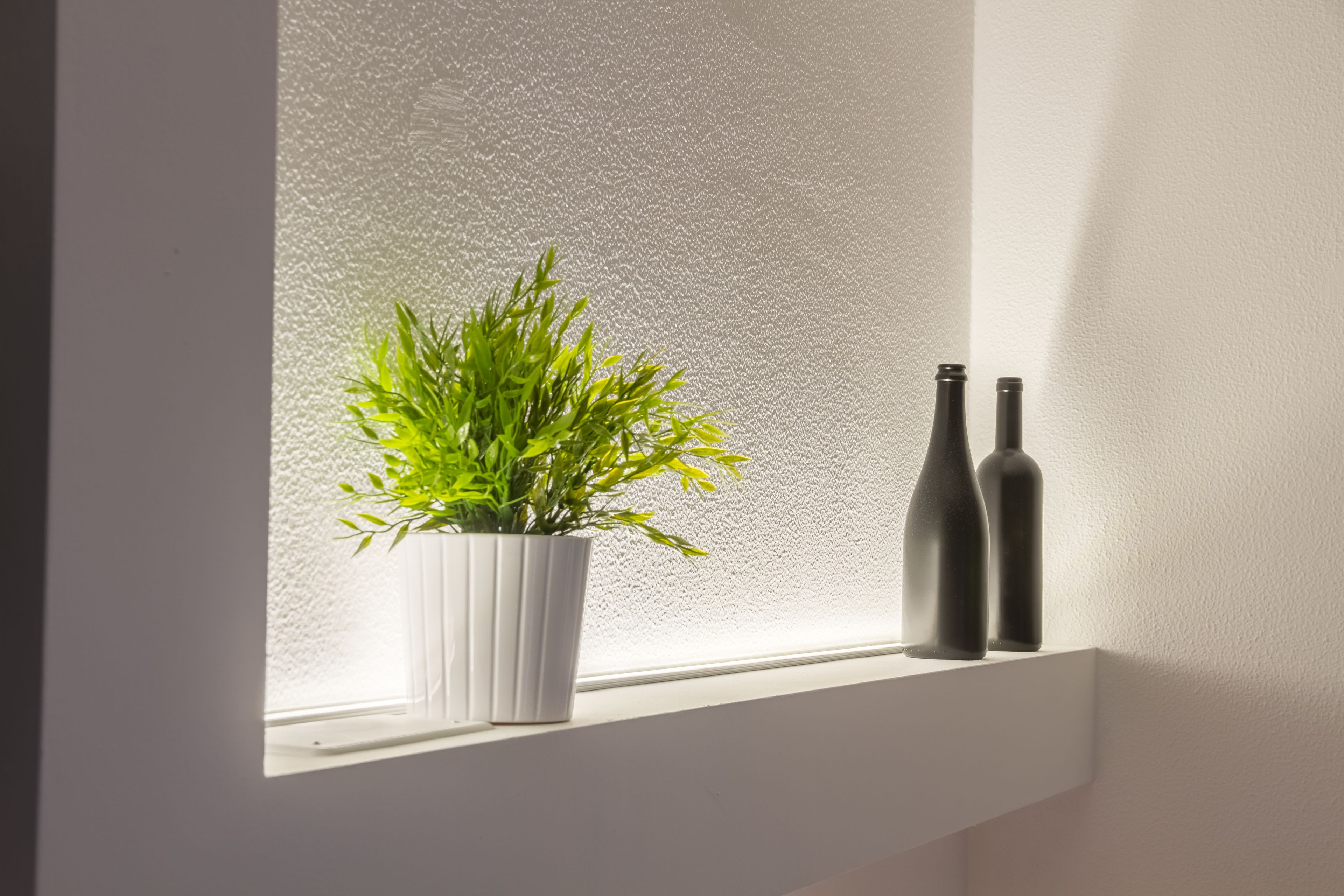 Un alféizar con una maceta blanca que contiene una planta verde, dos botellas oscuras y una pared con textura.