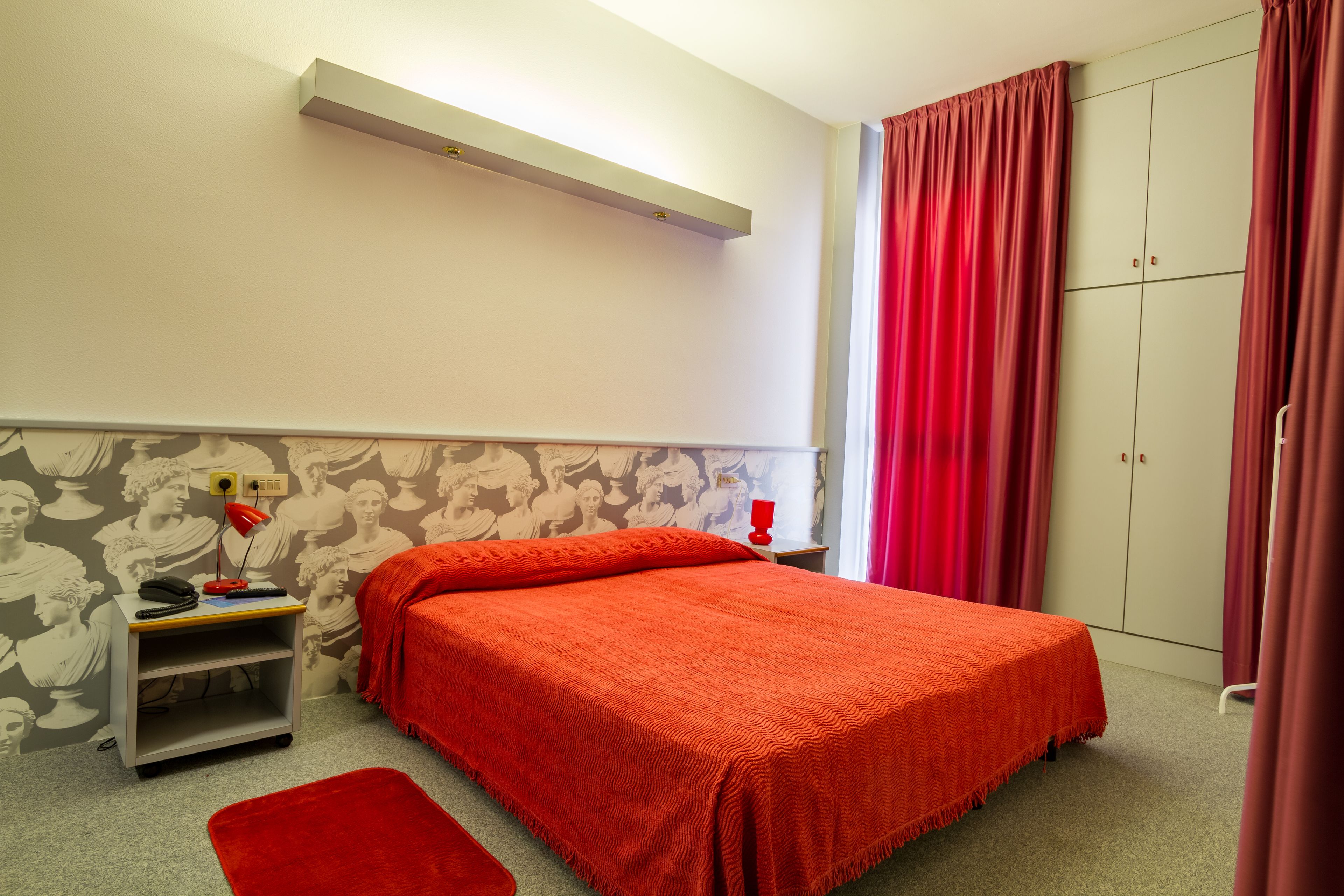 Un dormitorio con cama, mesita de noche, alfombra roja, armario y una pared con mural.